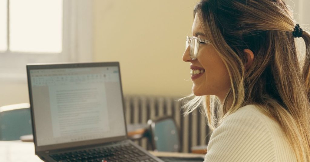 Smiling female teacher at desk with laptop in classroom setting during the day.